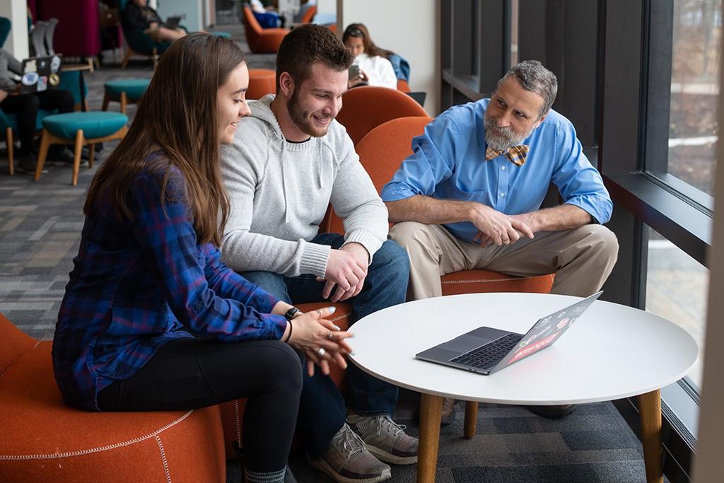一位大学教授和两位大学文科学生坐在一张配有笔记本电脑的桌子旁工作