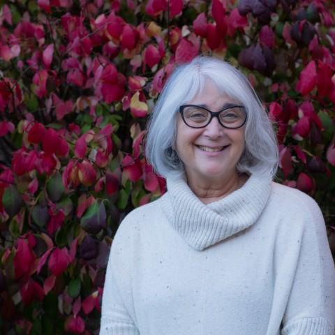 Photo of Shelley Cohen Konrad in white against a background of red leaves. 